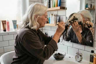 Femme aux cheveux blancs appliquant du henné avec un pinceau devant un miroir de salle de bain, bol de pâte henné et minuterie posés sur le plan de travail